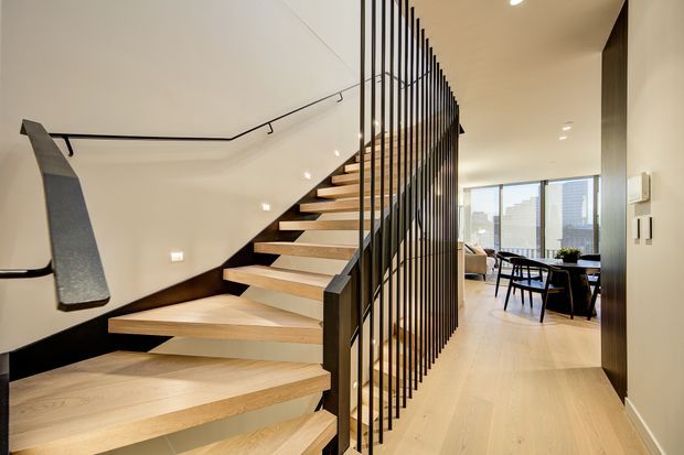 Interior view of a contemporary open-riser staircase with light timber treads, dark stringers, and a tall black metal vertical-bar balustrade. The stair hall opens toward a bright dining and living area with floor-to-ceiling glazing, pale timber flooring, recessed lighting, and dark trim details.