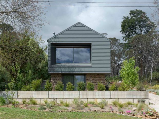 Front exterior view of a compact two-storey house set within landscaped gardens. The upper level has dark grey horizontal weatherboard cladding and a prominent recessed window, while the lower level combines grey cladding with exposed brown brick. A gabled roof, concrete boundary wall, native-style planting, and a gravel driveway are visible.