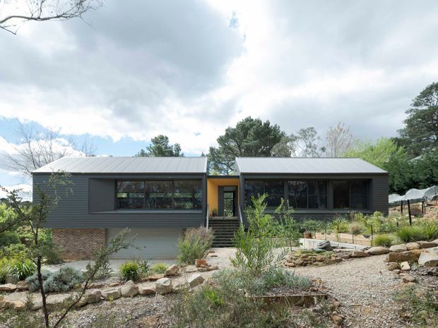 Single-storey contemporary residence composed of two dark-clad rectangular wings linked by a recessed central entry. The home has low-pitched metal roofs, broad dark-framed glazing, a brick lower level, and a landscaped front garden with gravel, native-style planting, and stone retaining edges.