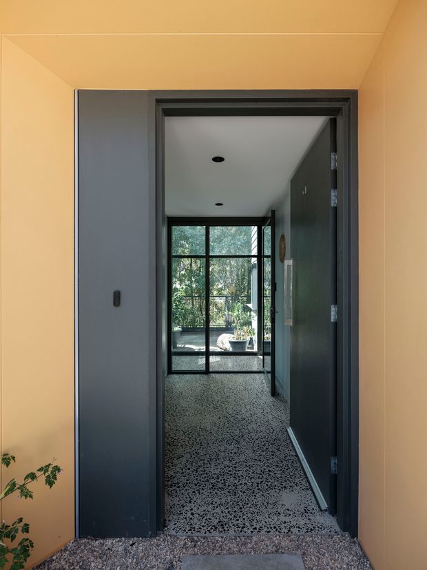 A narrow interior passage viewed through an open dark-grey door, leading toward a full-height black-framed glazed opening with leafy greenery visible beyond. The space has warm peach-painted walls and ceiling, a speckled terrazzo-style floor, dark charcoal doors and trim, recessed ceiling lights, and a small glimpse of a seating area at the far end. The foreground appears to transition from an outdoor or gravel-covered threshold into the hallway.
