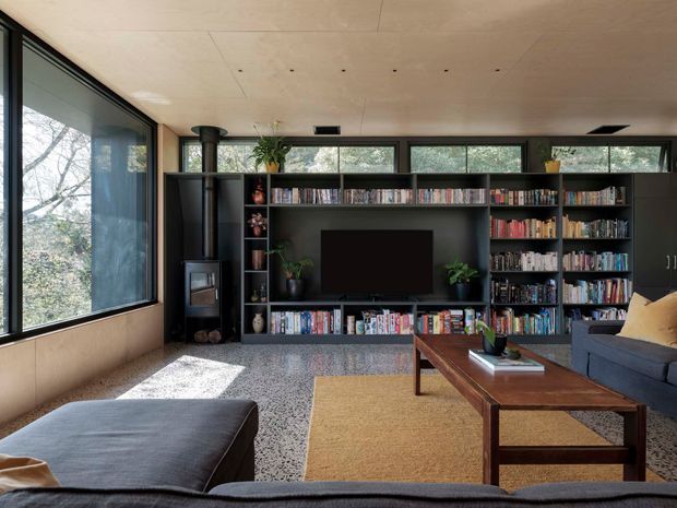 A warm, modern living room with expansive black-framed glazing, a full-height dark built-in entertainment and bookshelf wall, television, fireplace, large sectional sofa, upholstered ottoman, and a substantial timber coffee table. The room has a pale wood ceiling, textured grey flooring, layered rugs, and views of trees through the windows.