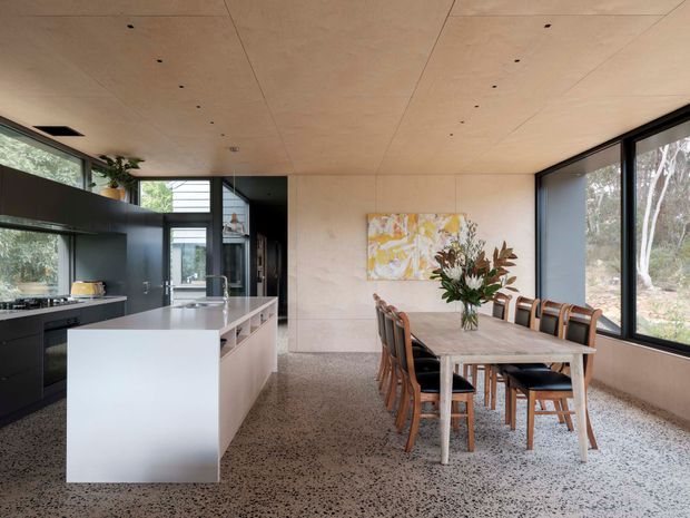 A contemporary open-plan kitchen and dining area with a long dining table beside large black-framed windows. The kitchen features dark built-in cabinetry, a pale island with an integrated sink, and a warm timber-panelled ceiling. Terrazzo-style flooring, natural light, and views of surrounding trees create a refined indoor-outdoor connection.