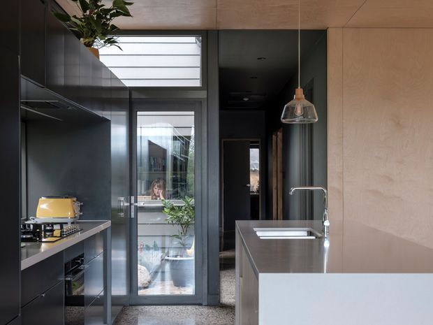 A compact contemporary kitchen viewed toward a glazed hallway and entry beyond. The kitchen features matte dark cabinetry, a slim stone or composite benchtop, integrated cooking appliances, a stainless-steel island with sink and tap, pale timber wall and ceiling lining, terrazzo-style flooring, and a sculptural pendant light. Full-height glazing and a clerestory bring natural light into the space.