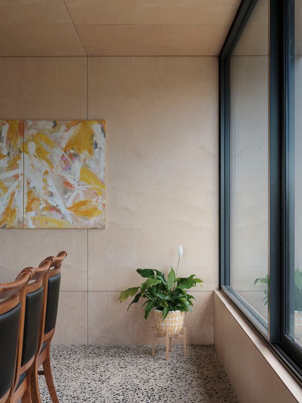 A refined dining area corner with full-height pale timber wall panelling, black-framed glazing, and a terrazzo floor. A row of sculptural wooden dining chairs is partially visible beside an abstract yellow, white, and orange artwork. A leafy plant sits on a small stand near the window, creating a warm, contemporary atmosphere.