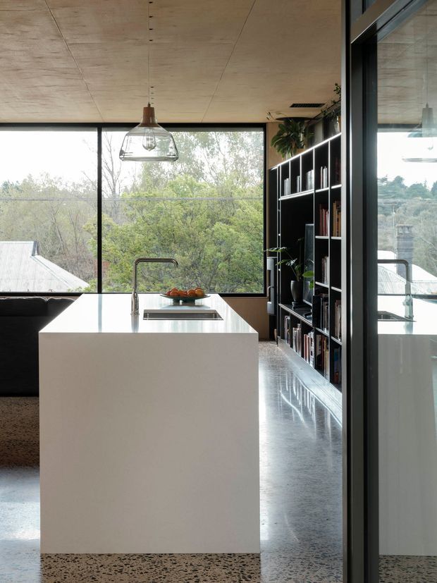 Contemporary open-plan kitchen with a large white island, integrated sink and cooktop, black-framed floor-to-ceiling glazing, a built-in black shelving wall, and a warm plywood ceiling. Terrazzo flooring and extensive views toward leafy surroundings create a refined, natural-feeling interior.