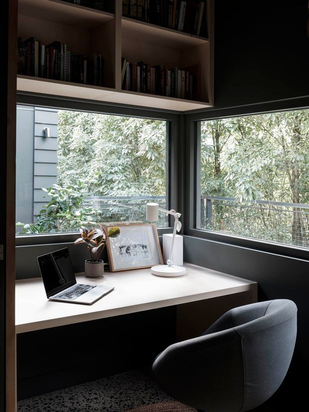 Compact, refined home office nook set into a corner with large windows overlooking dense greenery. A built-in dark desk with a pale worktop, laptop, desk lamp, plant and framed artwork sits beneath overhead open bookshelves. The room has dark walls and cabinetry, charcoal carpet, slim dark window frames, and a curved upholstered office chair, creating a calm, moody workspace with strong natural light.