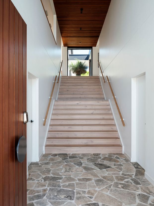 Bright residential entryway viewed from the front door, with a broad central staircase leading upward. The entry has white walls, warm timber stair treads and a timber-lined ceiling, slim timber handrails, a large dark timber pivot door, and an irregular natural stone floor. Glazing at the upper landing brings in daylight and reveals a planted courtyard element beyond.