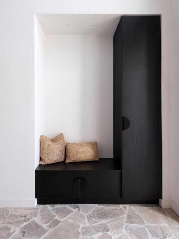 Minimal built-in entryway storage with a black timber-look tall cupboard, low bench cabinetry, and a recessed white alcove containing two woven neutral cushions. The floor is finished in irregular pale natural stone tiles with light grout.