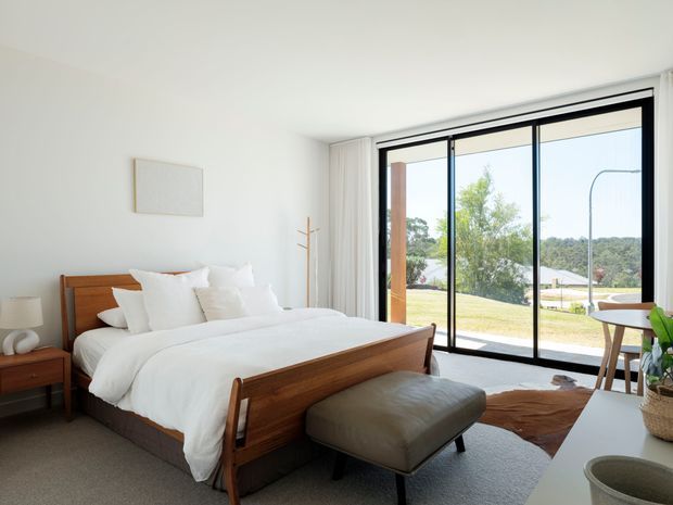Bright, minimalist bedroom with a low-profile timber bed, white bedding, upholstered bench, and small timber bedside table. Full-height black-framed glazed sliding doors open to a lawn and distant wooded landscape, creating an airy indoor-outdoor connection. A simple white wall artwork, freestanding coat rack, neutral carpet, and limited furnishings reinforce the restrained contemporary look.
