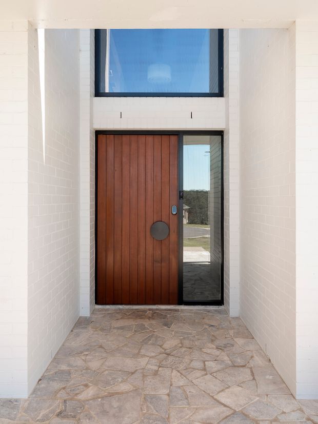 A covered residential entry porch viewed from outside, with white-painted brick walls, an irregular natural stone floor, and a tall timber pivot front door paired with a narrow glazed sidelight. A large horizontal transom window above the door brings in daylight, while dark-framed glazing and the door's vertical timber boards create a strong modern contrast.