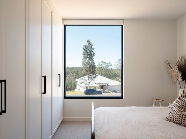 Bright, minimalist bedroom with a neatly made bed, full-height white built-in wardrobes with black handles, pale carpet, and a large black-framed window overlooking neighbouring homes and greenery. Neutral finishes and simple dried botanical decor create a calm, contemporary feel.