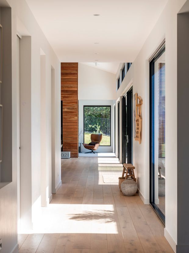 A bright, elongated residential hallway with warm timber flooring, white walls and ceiling, recessed downlights, and extensive black-framed glazing along the right side. A tall vertical timber feature panel anchors the far end, where a lounge chair sits beside a garden-facing window. Natural light creates strong rectangular shadows across the floor, with a small stool, woven basket, and wall-mounted decorative timber element adding warmth.
