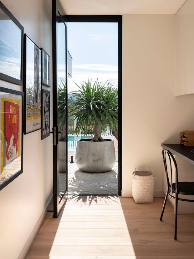 Bright, narrow interior workspace or study area opening through a full-height black-framed glazed door to an outdoor terrace and swimming pool. The room has pale timber flooring, warm off-white walls, framed artwork, a compact dark desk with a black chair, and a woven wastebasket. A large potted plant and pool are visible outside, with strong sunlight casting shadows across the floor.