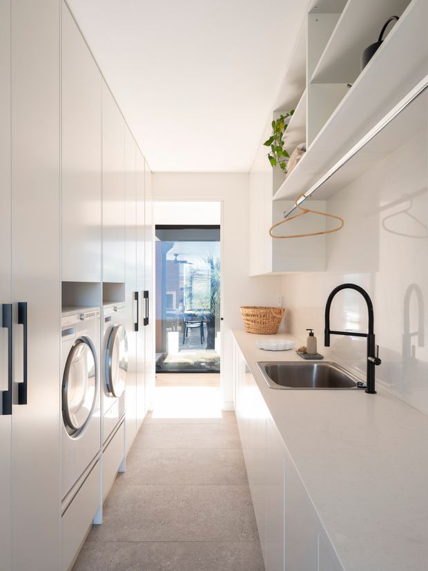 Bright, narrow contemporary laundry room with full-height white cabinetry, integrated front-loading washer and dryer, a long white benchtop, stainless-steel undermount sink, and a black gooseneck tap. Open shelving with hanging rail, woven basket and indoor plant add practical storage and warmth. A glazed door at the end brings in strong natural light and provides a view to an outdoor area.