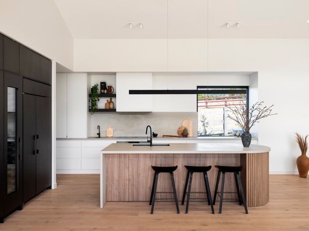 Bright contemporary kitchen with a large central island, integrated white cabinetry, dark full-height pantry or appliance joinery, timber flooring, and a warm timber island front. Three black stools sit at the island, which includes a pale stone benchtop and black undermount sink tapware. A wide window brings in natural light, while open shelving and restrained decorative branches add subtle styling.