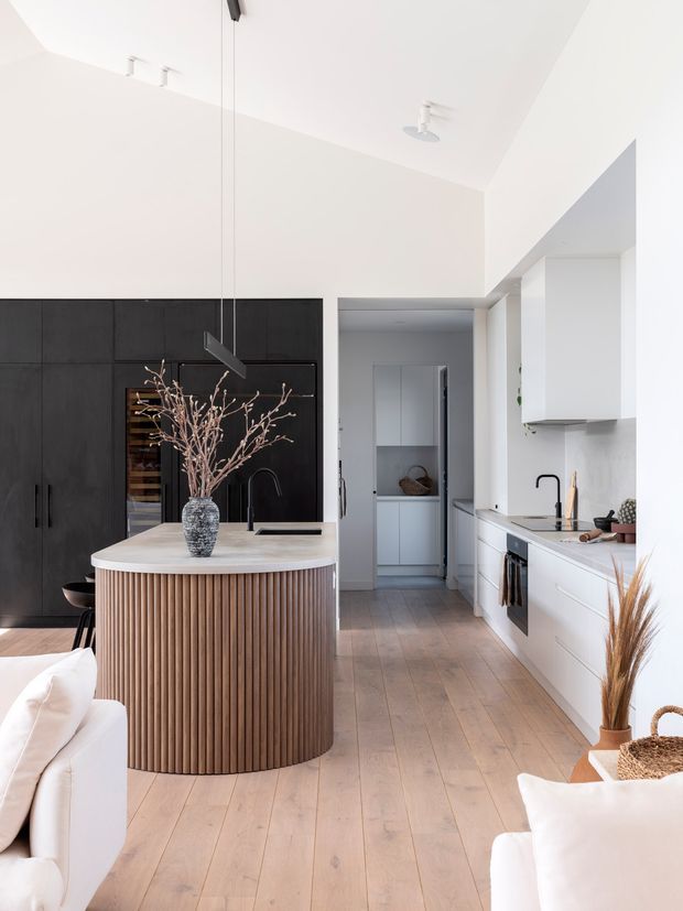 A bright contemporary kitchen viewed from an adjoining living area, featuring a curved island with vertically fluted timber cabinetry, a pale stone benchtop, matte black tall cabinetry and integrated appliances, white perimeter cabinetry, black tapware, and light timber flooring. A compact pantry or utility space is visible beyond the kitchen.