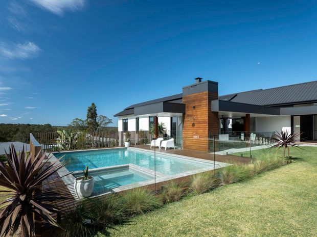 Exterior view of a modern single-storey residence with dark pitched metal roofing, white rendered walls, extensive glazing, and a prominent timber-clad chimney or feature wall. The landscaped backyard includes a swimming pool with glass balustrades, ornamental grasses, and sculptural planting, overlooking a rural or bush setting.