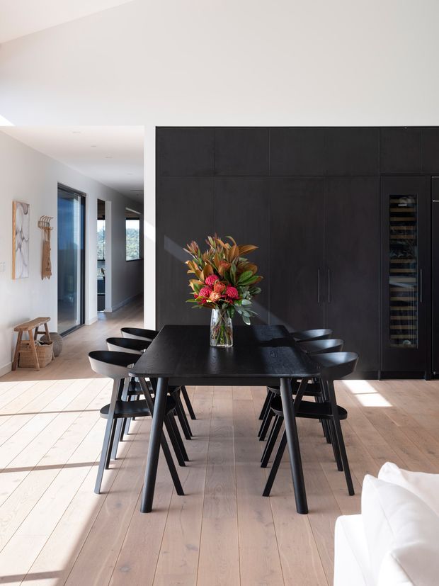 Bright contemporary open-plan dining area with a large black dining table and matching chairs, positioned in front of extensive dark built-in cabinetry that includes a tall integrated wine refrigerator. Pale timber flooring, white walls, expansive glazing, and a partially visible white sofa create a clean, light-filled interior. A floral arrangement forms the centrepiece of the table, while a hallway and small seating area extend into the background.