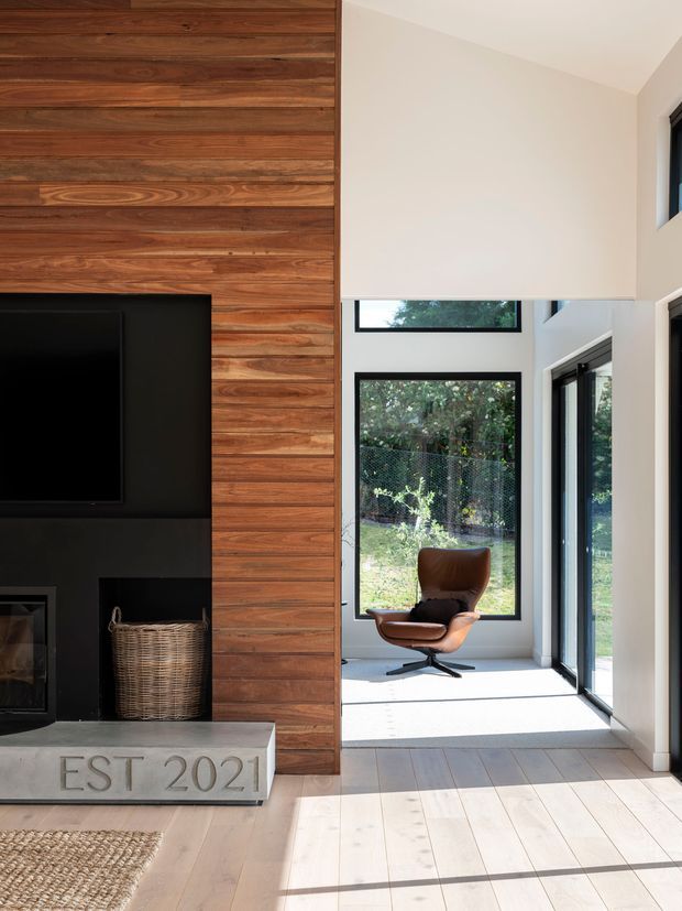 Bright contemporary living area with a double-height corner of floor-to-ceiling glazing, a black built-in media and fireplace wall, and a prominent vertical feature wall clad in warm horizontal timber boards. Pale timber flooring, white walls, and a brown leather lounge chair create a clean, warm interior. A small fireplace basket and textured rug are also visible.