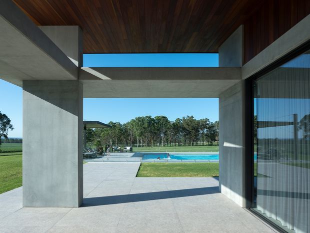 A covered outdoor terrace or passage framed by large raw concrete walls and beams, with a dark timber-lined ceiling and extensive glazing on the right. The opening looks onto a swimming pool, paved pool terrace, lawn and rural tree-lined landscape. Strong geometric composition, deep shadows and bright blue sky create a modern indoor-outdoor connection.