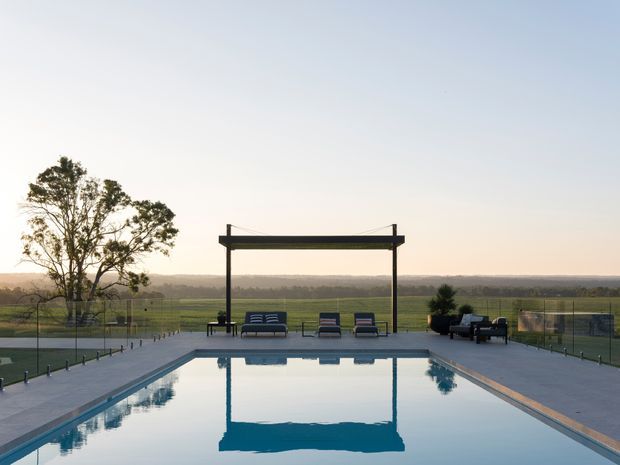 A rectangular outdoor swimming pool occupies the foreground, reflecting a simple dark overhead shade structure. Poolside loungers and seating are arranged on a paved terrace, with transparent fencing and open rural farmland stretching into the distance. The scene has a restrained, contemporary outdoor design and soft evening light.