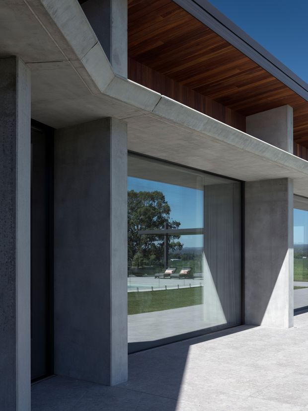 Close-up exterior view of a highly modern concrete residence, showing strong exposed concrete columns, deep projecting slab edges, full-height glazed openings, and a warm timber-lined recessed soffit or upper-level ceiling. The glazing reflects a landscaped yard and swimming pool, while the composition emphasizes the building’s heavy structural geometry and material contrast.