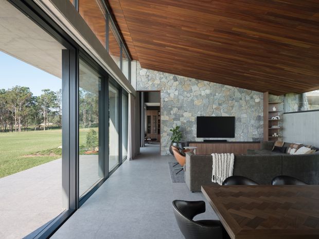 A contemporary open-plan living and dining space with full-height black-framed glazing opening onto a lawn, a warm timber-lined ceiling, a natural irregular-stone feature wall, polished concrete flooring, and dark timber dining furniture. A television and built-in shelving define the living area, while the dining table occupies the foreground.