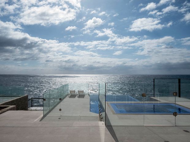 A contemporary seaside terrace viewed from above, featuring a rectangular swimming pool, clear glass balustrades, pale paved surfaces, and lounge chairs overlooking the ocean. The expansive horizon, reflective water, and dramatic partly cloudy sky create a luxurious coastal setting.