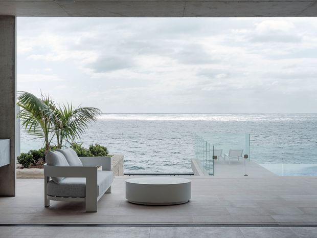 A minimalist coastal outdoor living area opens directly onto an ocean view. The covered terrace features pale large-format tiled or stone-like flooring, a grey upholstered lounge chair, a low round table, and a glass balustrade around a pale blue pool or water feature. A palm and low planting soften the otherwise clean, concrete-toned setting.