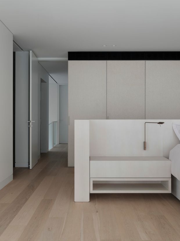 A minimalist bedroom viewed toward a built-in bedhead and bedside unit. The room uses pale timber flooring, white and light-grey built-in cabinetry, flush doors, and a restrained linear wall-mounted reading light. A hallway and open door are visible to the left, while the bed is partially visible on the right. The overall composition is clean, quiet, and highly streamlined.