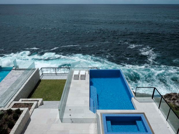 A modern coastal exterior terrace overlooking the ocean, with a vivid blue rectangular swimming pool and integrated spa, pale stone-paved surfaces, frameless glass balustrades, landscaped lawn and raised planters. Waves break directly below the elevated deck, creating a dramatic oceanfront setting.