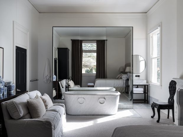 Elegant, light-filled traditional bathroom with a freestanding sculptural bathtub positioned in front of a large wall mirror. The room includes a pale upholstered sofa, dark timber doors and cabinetry, a small black side table, classic drapery, detailed ceiling cornices, and a glimpse of a bedroom reflected in the mirror. The palette is predominantly white, soft grey, and black, creating a refined, luxurious atmosphere.