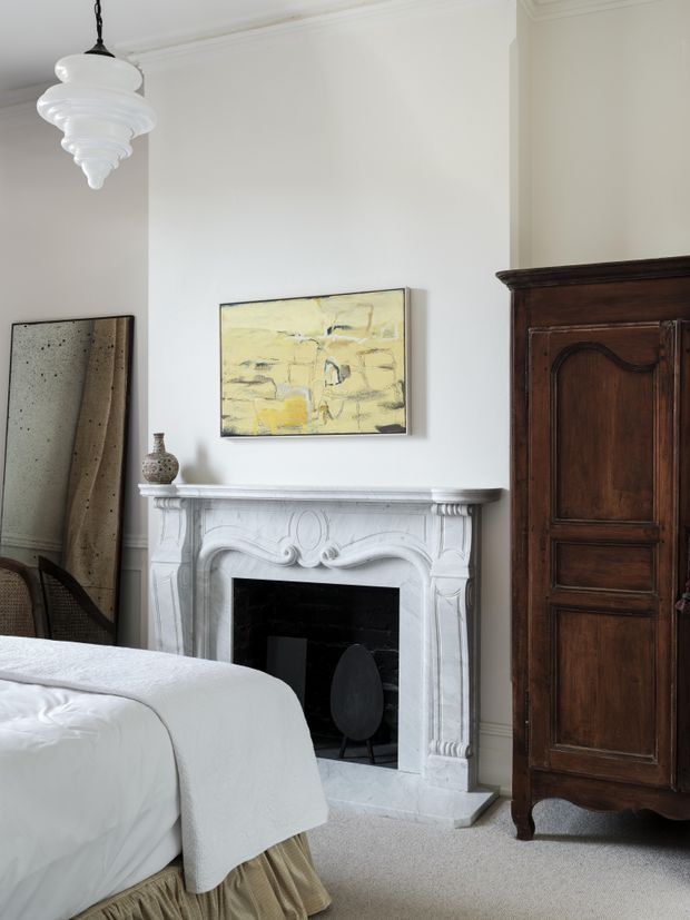A refined bedroom interior with a white upholstered bed in the foreground, an ornate carved white marble or stone fireplace, pale walls with decorative cornice moulding, a dark antique timber armoire, and a sculptural white pendant light. A muted abstract artwork and small decorative vessel sit above and beside the fireplace, creating a calm, traditional yet softly eclectic atmosphere.