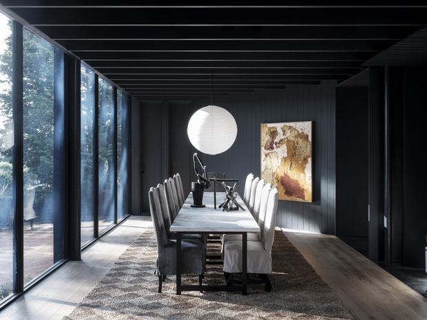 A dramatic, dark-toned dining room with a long rectangular dining table and upholstered chairs for a large group. Full-height glazed panels line the left wall, bringing in views of trees and strong natural light. The room features dark vertical wall panelling, a black exposed-beam ceiling, timber flooring, a large textured rug, a spherical pendant light, and a warm-toned abstract artwork.
