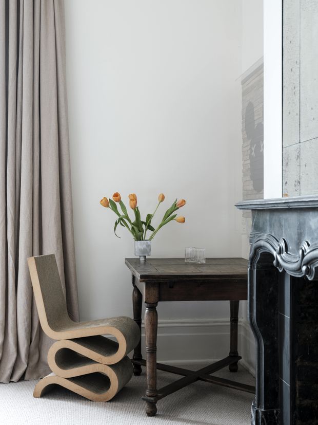 A refined interior corner featuring a sculptural curved wooden lounge chair beside a dark antique-style timber side table with turned legs and a lower stretcher. A vase of orange tulips sits on the table. Full-height taupe curtains, pale walls, light carpet, and an ornate dark marble fireplace create a quiet, elegant setting.