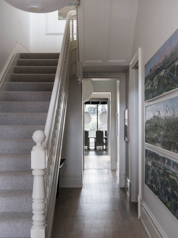 A refined residential hallway with a carpeted staircase and detailed white painted balustrade on the left. The narrow corridor has pale grey-white paneled walls, decorative trim, timber flooring, framed artwork, and a view through to a bright dining area with large glazing and pendant lights. The space has a traditional, elegant character with soft neutral tones.