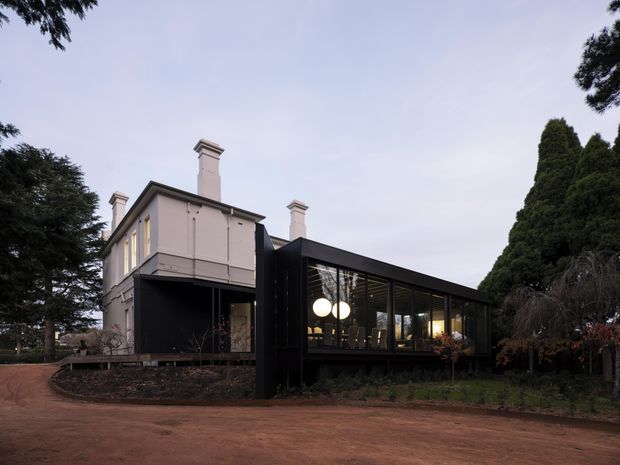 Exterior view of a traditional two-storey rendered house extended by a long, dark, glazed contemporary pavilion. The original house has a pale facade, tall chimneys and classical proportions, while the addition features black cladding, extensive floor-to-ceiling glazing and warm interior lighting. Mature trees and a broad unsealed driveway surround the property.