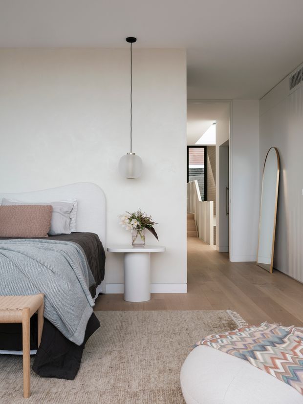 A serene contemporary bedroom with a low upholstered bed, layered neutral and blue-grey bedding, a small cylindrical bedside table, pendant light, and floral arrangement. Pale timber flooring, a textured neutral rug, white walls, and a full-length arched mirror create a bright, refined interior. A hallway and stair area are visible beyond the bedroom doorway.