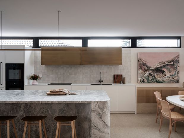 Contemporary kitchen and dining area with a large veined stone island, timber cabinetry, pale textured tiled splashback, integrated dark appliances, and a long horizontal clerestory window band. A round dining table and woven timber chairs sit beside built-in timber joinery and a large abstract artwork.
