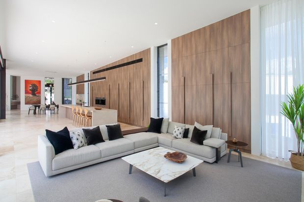 A bright, spacious contemporary living room with a large L-shaped light-grey sectional sofa, black and patterned cushions, and a marble-look rectangular coffee table. The room features full-height warm timber wall panelling with integrated linear detailing and a fireplace, pale flooring, recessed ceiling lights, floor-to-ceiling sheer curtains, and a nearby open-plan kitchen and dining area. A tall indoor plant and minimal furnishings add a refined, airy atmosphere.