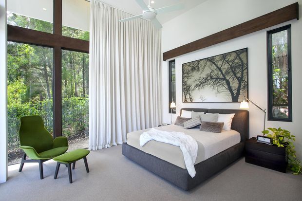 A bright, contemporary bedroom with a large upholstered bed, layered neutral bedding, dark timber architectural beams, floor-to-ceiling glazing, sheer curtains, and a green lounge chair with ottoman. The room opens visually to a leafy wooded setting and features a large monochrome tree artwork above the bed, slim bedside tables, and modern pendant and floor lighting.
