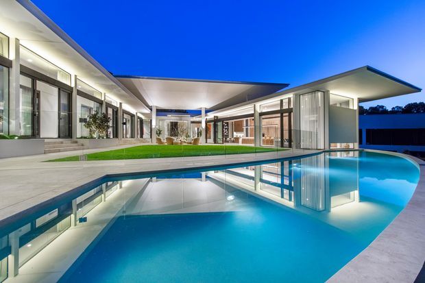 Twilight exterior view of a large contemporary residence arranged around a swimming pool. The composition features crisp flat roof planes, extensive floor-to-ceiling glazing, white rendered walls, concrete paving, illuminated interiors, and strong reflections across the pool surface.