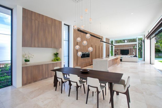 Bright open-plan contemporary interior combining a dining area, kitchen and living room. A large dark timber dining table with light upholstered chairs sits in the foreground, while timber cabinetry, a white island, marble-look flooring, sculptural pendant lighting and extensive glazing create an airy, refined space. The living area and garden are visible beyond the kitchen.