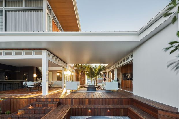 A sophisticated open-air courtyard and entertaining terrace beneath a large cantilevered flat roof. The space features extensive timber decking and stepped timber platforms, white rendered walls, timber-lined soffits, clerestory-style ventilation openings, built-in seating, an outdoor dining area, and a central palm-planted feature. Interior living and kitchen areas are visible around the courtyard, creating a seamless indoor-outdoor connection.