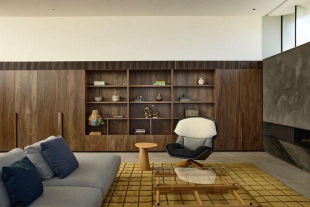 A refined mid-century-inspired living room with a large grey sectional sofa, dark timber built-in cabinetry and open shelving, a sculptural lounge chair, nested timber coffee tables, and a muted mustard grid-pattern rug. A dark stone or concrete fireplace wall and high clerestory glazing add architectural interest. The room has a warm, understated and curated appearance.