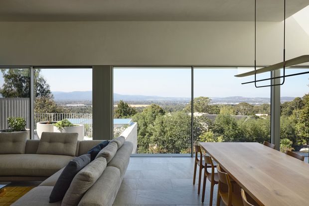 A contemporary open-plan living and dining area with expansive floor-to-ceiling glazing overlooking a leafy suburban landscape and distant hills. The room features a large neutral sectional sofa, a long timber dining table with chairs, pale tiled flooring, a restrained neutral palette, and a sculptural linear pendant light. The space is bright, refined, and strongly connected to the outdoor view.