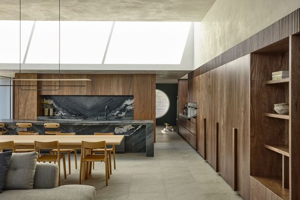 A refined open-plan kitchen and dining area with extensive dark timber cabinetry, a dramatic veined stone island and splashback, a long timber dining table, and an integrated living area in the foreground. A broad skylight brings bright natural light into the space, while a textured concrete ceiling and large-format grey flooring reinforce the contemporary architectural character.