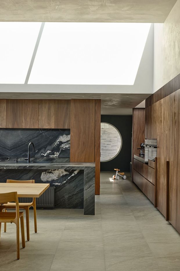 A sophisticated open-plan kitchen and dining area with extensive dark walnut cabinetry, a dramatic black-and-white veined stone island and splashback, pale large-format tiled flooring, and a concrete-textured ceiling. A large rectangular skylight floods the space with natural light. The kitchen opens toward a dark hallway with a round window or glazed opening, while a simple timber dining table and chairs occupy the foreground.