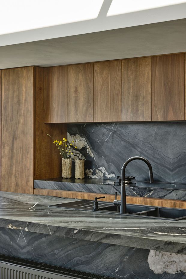 Close-up view of a sophisticated contemporary kitchen featuring extensive dark natural-stone surfaces across the island benchtop, waterfall edge and splashback, paired with warm timber cabinetry. A black undermount sink and tall arched tap are prominent in the foreground, while integrated upper cabinets and subtle ceiling detailing complete the composition.