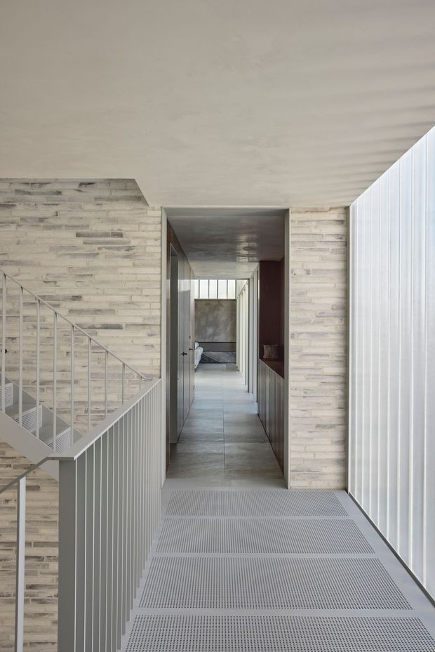 A minimalist architectural hallway and stair landing with pale exposed masonry walls, a grey tiled corridor floor, metal stair balustrades, a perforated metal landing floor, and translucent vertical glazing along the right side. The corridor leads toward a small seating or lounge area, while the restrained palette and strong linear composition create a quiet contemporary atmosphere.